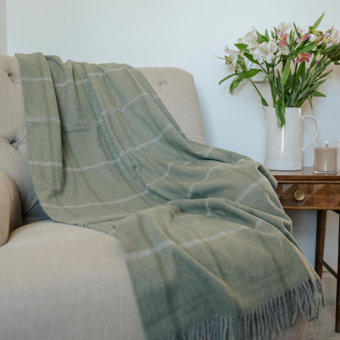 The Eskdale meadow sage green pure cashmere throw blanket with a pale grey and green windowpane check pattern and fringed edges draped over a beige armchair in a cosy living room setting.