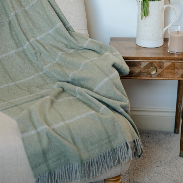 A sage green plaid throw blanket with white windowpane lines and fringed edges draped over a light beige armchair next to a wooden side table.