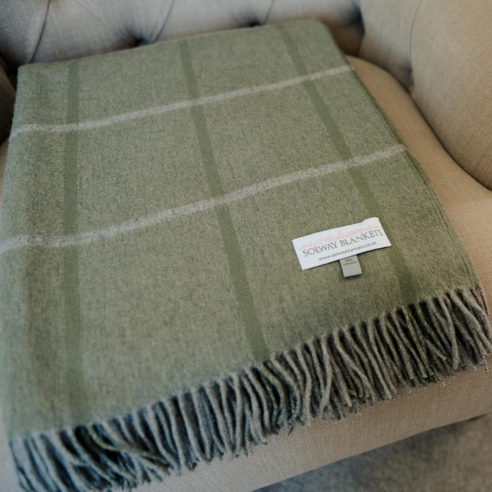 Eskdale Meadow pure cashmere throw in soft sage green with delicate windowpane check and twisted fringe folded neatly on an upholstered armchair. A white brand label reading 'SOLWAY BLANKETS' is visible on the blanket.
