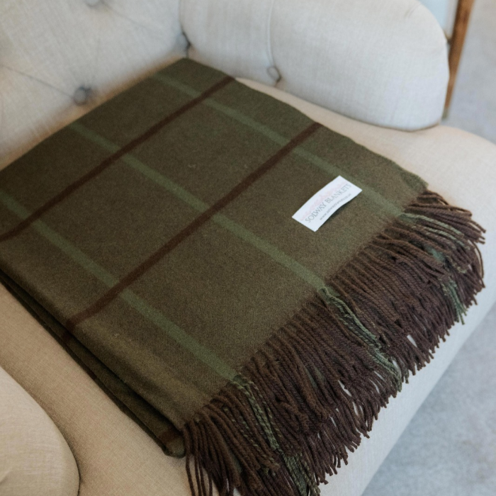 A moss green cashmere throw with brown and green windowpane overcheck and fringe, folded on a light beige upholstered armchair. A small white woven label with 'SOLWAY BLANKETS' is visible on the blanket.
