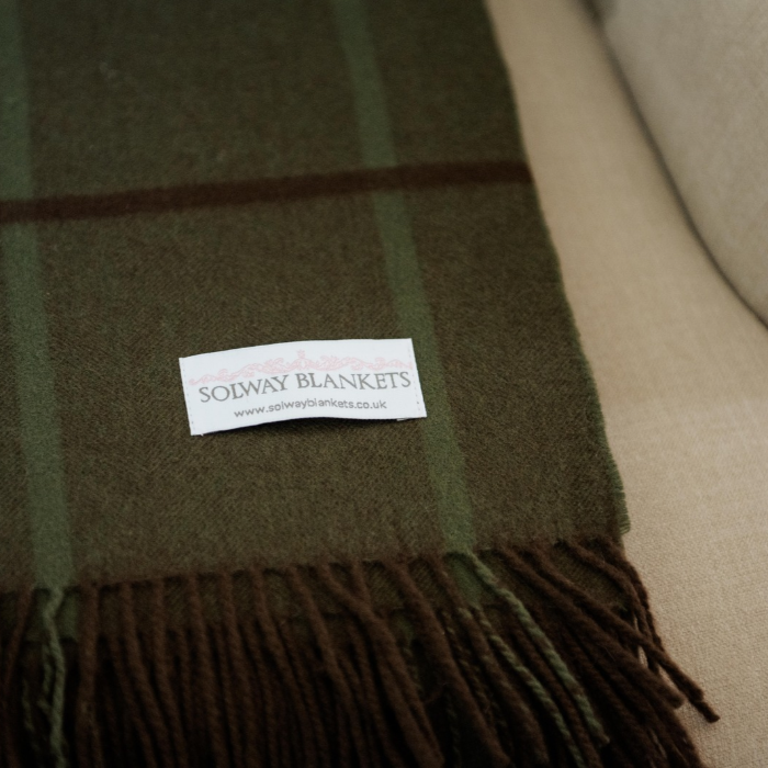 A close-up of the Eskdale moss green pur cashmere throw with brown and mid green over check and delicate fringe, featuring a white label that reads 'SOLWAY BLANKETS'.