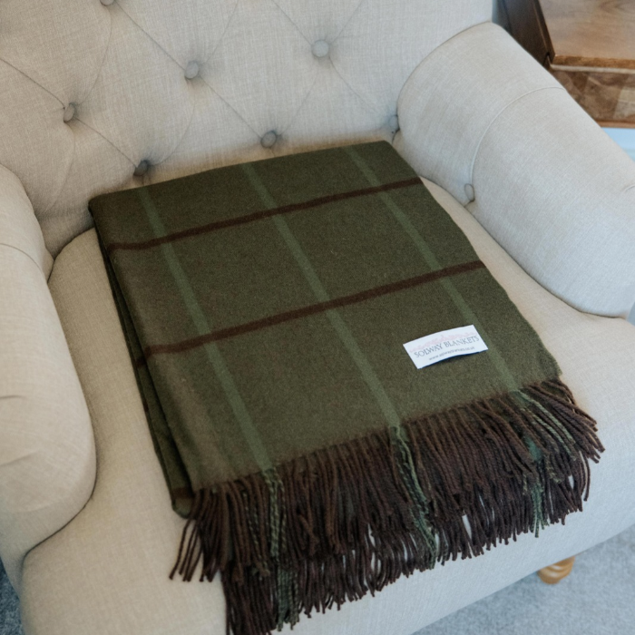 An moss green with brown over check, pure cashmere wool throw blanket with and fringe finish, displayed on a beige tufted armchair. A small white woven label is visible on the corner of the blanket.