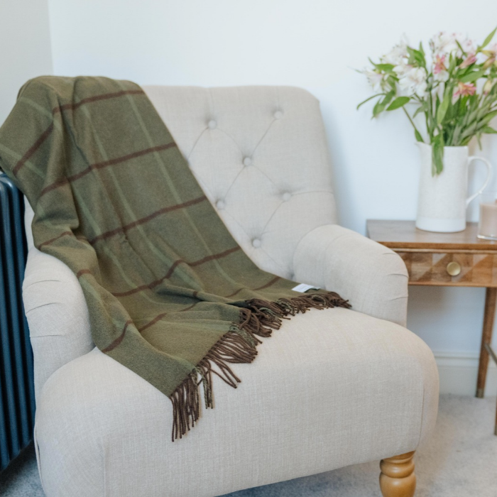 An moss green cashmere windowpane check throw blanket with dark brown and olive green and a twisted fringe is draped over a light beige tufted armchair in a styled room setting.