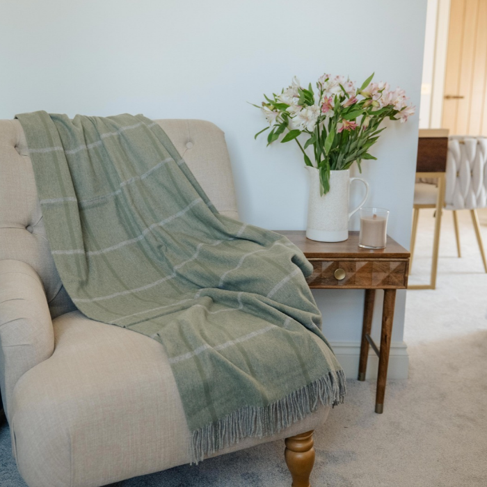 The Eskdale sage green plaid cashmere throw blanket with fringe is draped over a beige armchair in a bright, modern living room next to a wooden side table holding a vase of pink and white flowers.