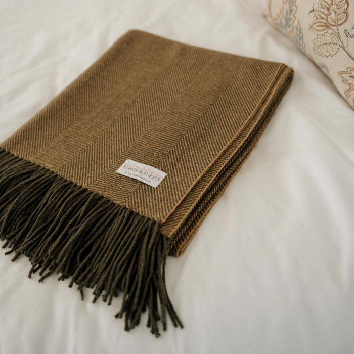 A folded Olive-grey and ochre herringbone throw blanket with dark fringe and a small white 'SOLWAY BLANKETS' label, placed on a white bed next to a patterned pillow.