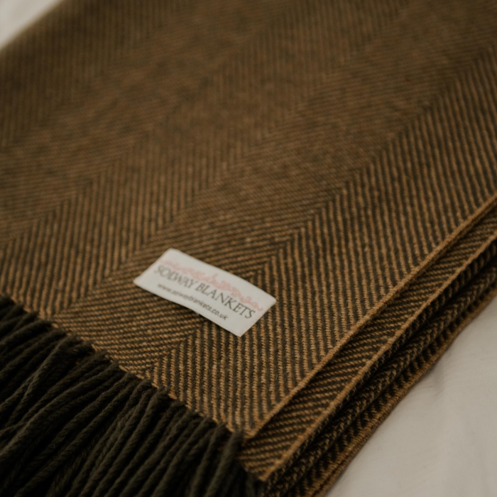 A close-up of a folded herringbone wool blanket in a warm ochre brown colour, featuring a white woven label that reads 'SOLWAY BLANKETS' and olive-grey fringe along one edge.