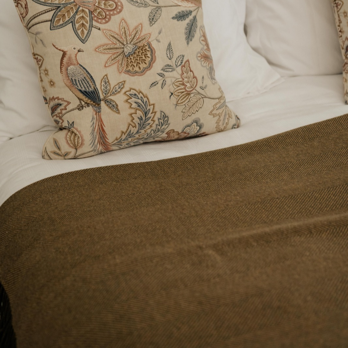 A close-up of a textured Olive-grey and ochre herringbone lambswool blanket draped over white bedding, with a decorative beige pillow featuring a bird and floral pattern resting on top.