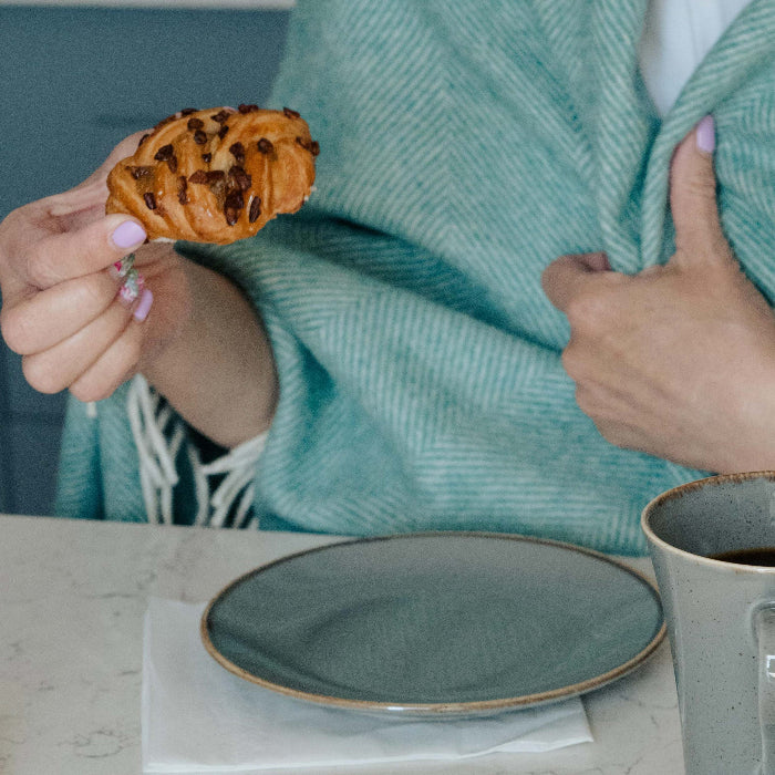 Person holding a pastry and a mug on a kitchen counter wrapped in a cosy eucalyptus herringbone throw