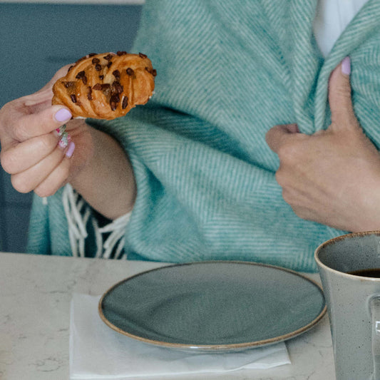 Person holding a pastry and a mug on a kitchen counter wrapped in a cosy eucalyptus herringbone throw