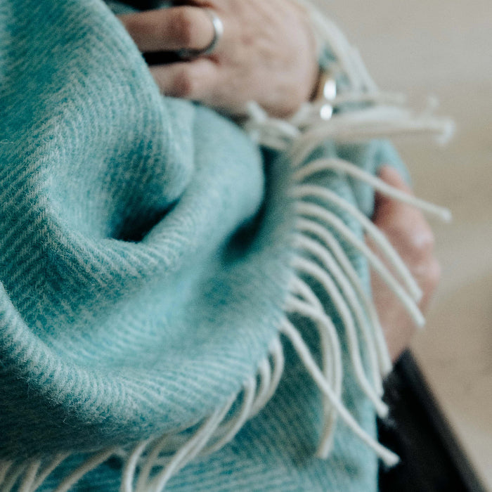 Close up of a teal coloured herringbone throw with white fringe wrapped around a person. 