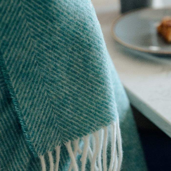 Up close focused picture of a teal coloured herringbone blanket throw with white fringe draped beside a marble surface, with a blurred plate of food in the background.