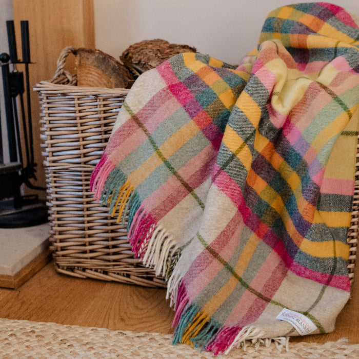 Falmouth Pink & Ivory Check Shetland Wool throw a Multicolored plaid blanket draped over a wicker basket on a wooden floor.