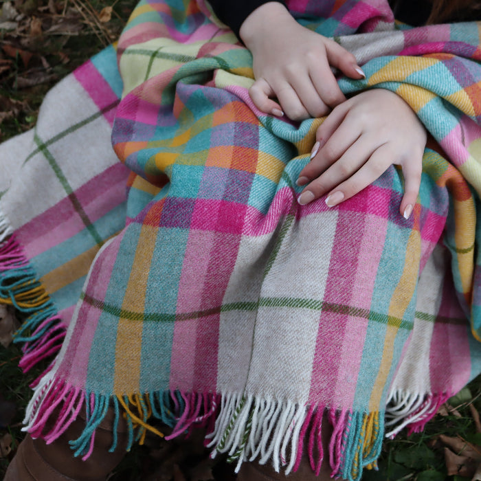 Person wrapped in the Falmouth Pink & Ivory Check Shetland Wool Blanket a colourful plaid throw sitting on the ground.