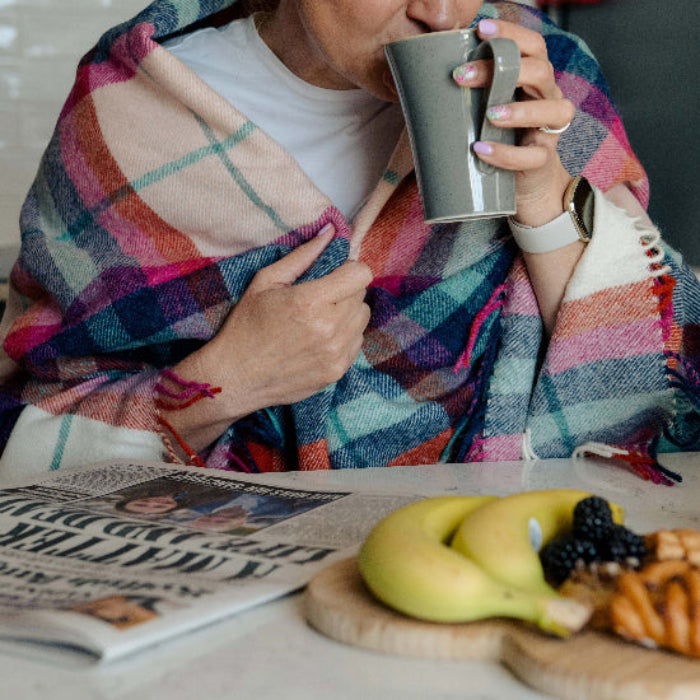 Person wrapped in a colourful Falmouth Red & Ivory Check Shetland Wool Blanket drinking from a mug with a newspaper and fruit on the table.