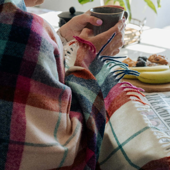 Person sitting on a stool wrapped with a Falmouth Red & Ivory Check Shetland Wool Blanket, holding a mug and looking at food on a tray and a newspaper