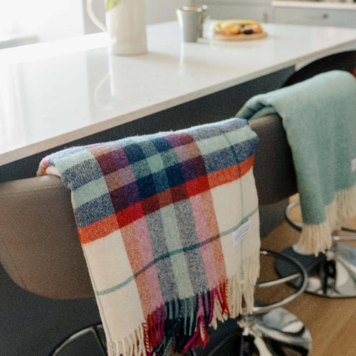 Falmouth Red & Ivory Check Shetland Wool Blanket a colourful plaid blanket draped over a bar stool in a kitchen setting with a vase of tulips on the counter.