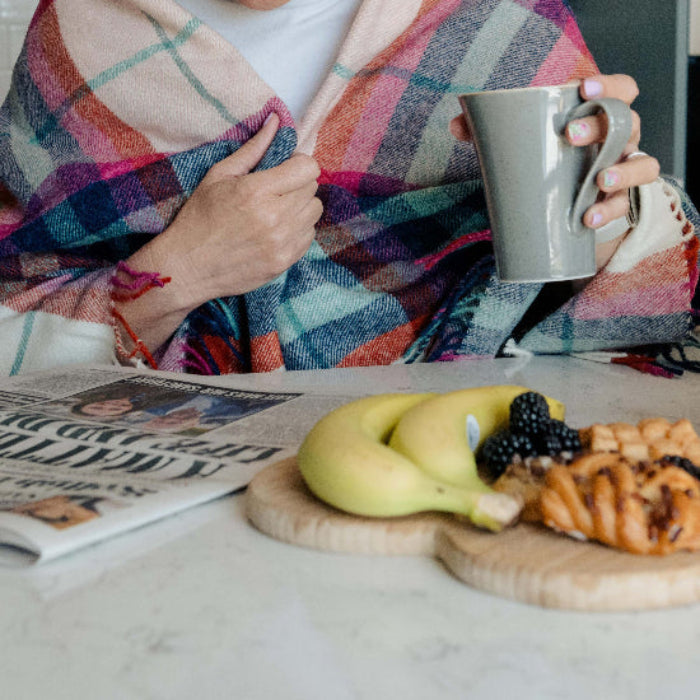 Person wrapped up in the Falmouth Red & Ivory Check Shetland Wool Blanket holding a mug with breakfast items and a newspaper on a table