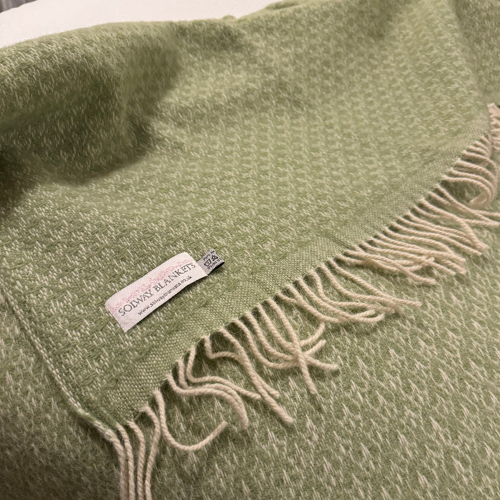 Fern green wool throw with fringes and a Solway Blankets label on a white bed