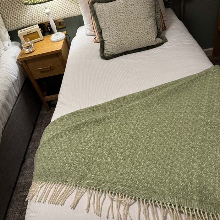 Fern green wool throw with fringe draped over bed with striped and spotted cushions in a bedroom setting