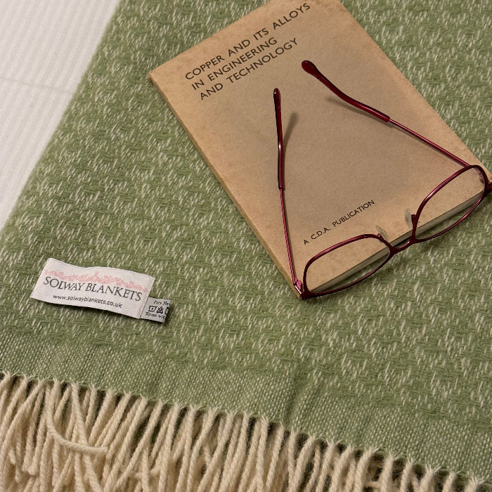 Fern green wool throw with fringe and a visable Solway Blankets label with a pair of glasses and a book placed on top 