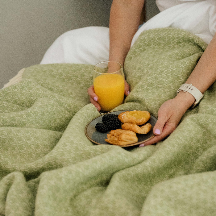 Person holding a glass of orange juice and a plate of pastries under a cosy fern green wool throw.