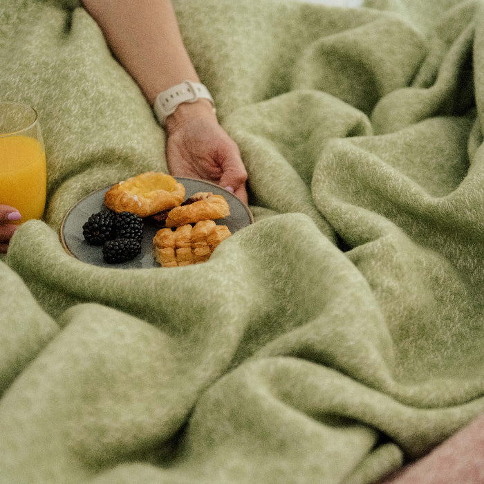 Person holding a glass of orange juice and a plate of pastries under a fern green wool blanket.