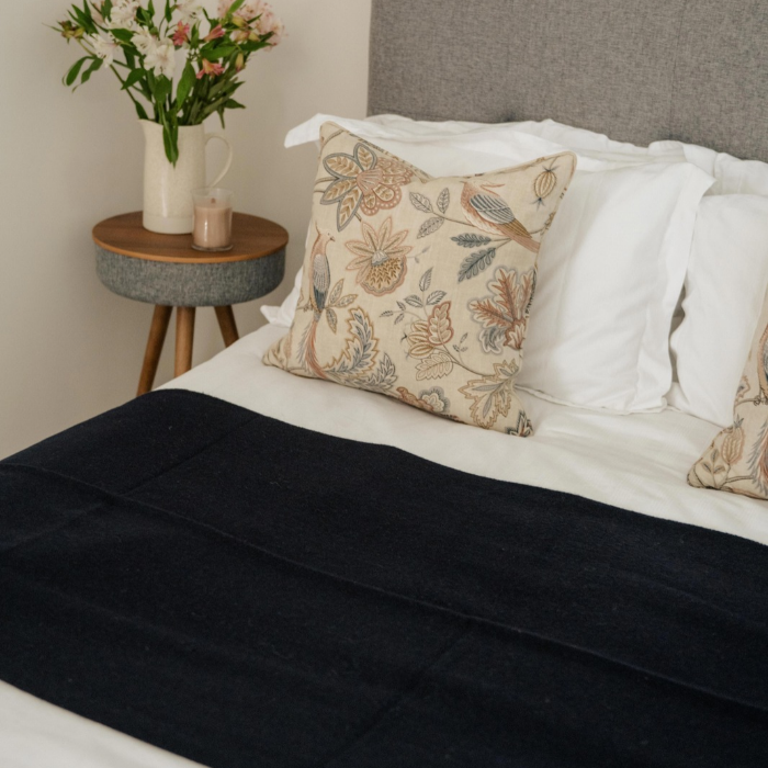 A styled bedroom scene showing a bed with white bedding, Moffat Midnight pure cashmere throw in deep navy blue with fine weave and a beige floral accent pillow. A small round side table with a vase of flowers and a candle sits beside the bed.
