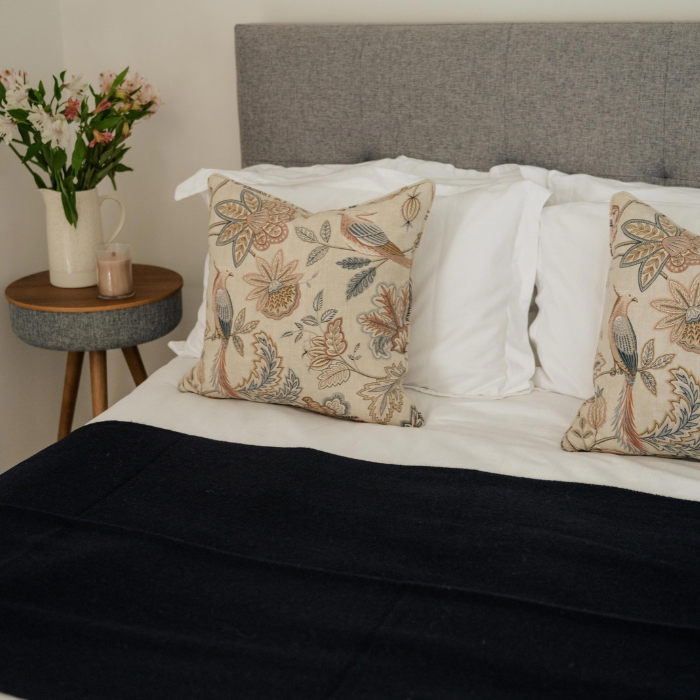 A styled bedroom scene showing a bed with white bedding, a dark navy pure cashmere Scottish woven blanket, and two decorative throw pillows with a beige floral and bird pattern. A grey upholstered headboard and a small wooden side table with flowers are visible.