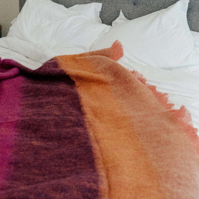 Bed with the Fuchsia Mohair Throw a striped blanket featuring purple and orange hues, white pillows, and a gray headboard.