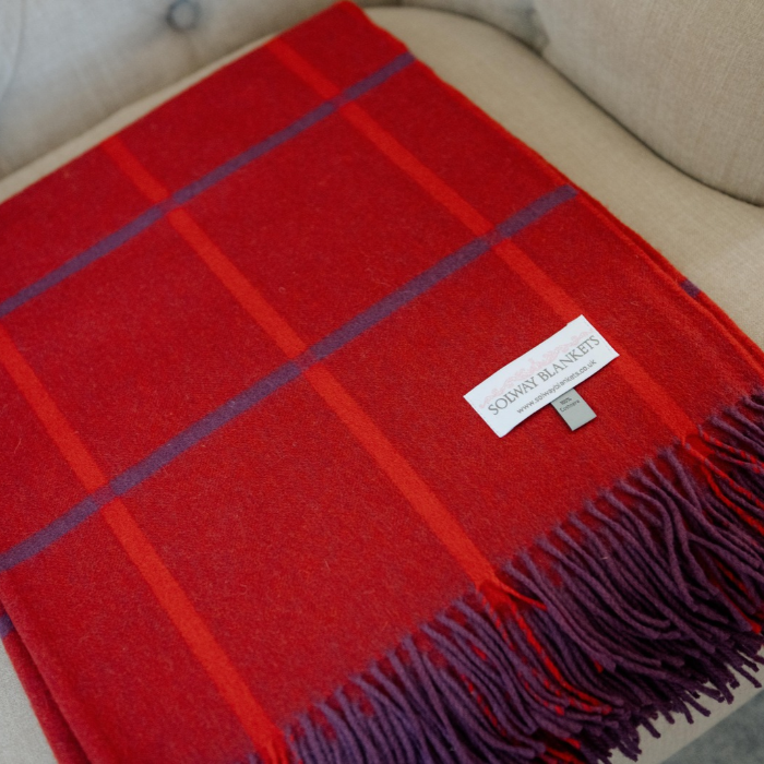 Close-up of a folded red and purple windowpane check throw, woven in Scottish in lure luxury cashmere with fringed edges, displayed on a beige upholstered armchair. A white brand label reading 'SOLWAY BLANKETS' is visible.