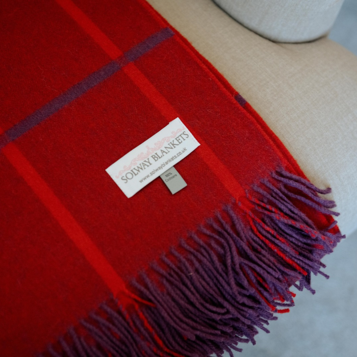 A close-up of a red plaid wool blanket with purple fringe and stripes, laid on a beige upholstered chair. A white label reads 'SOLWAY BLANKETS'.