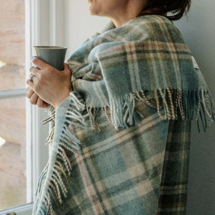 Person wrapped in the Glen Coe Shetland Pure New Wool Throw a plaid blanket, holding a mug by a window