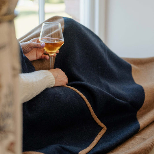Person holding a glass of wine wrapped in a navy and tan cashmere blanket throw, sitting by a window. 