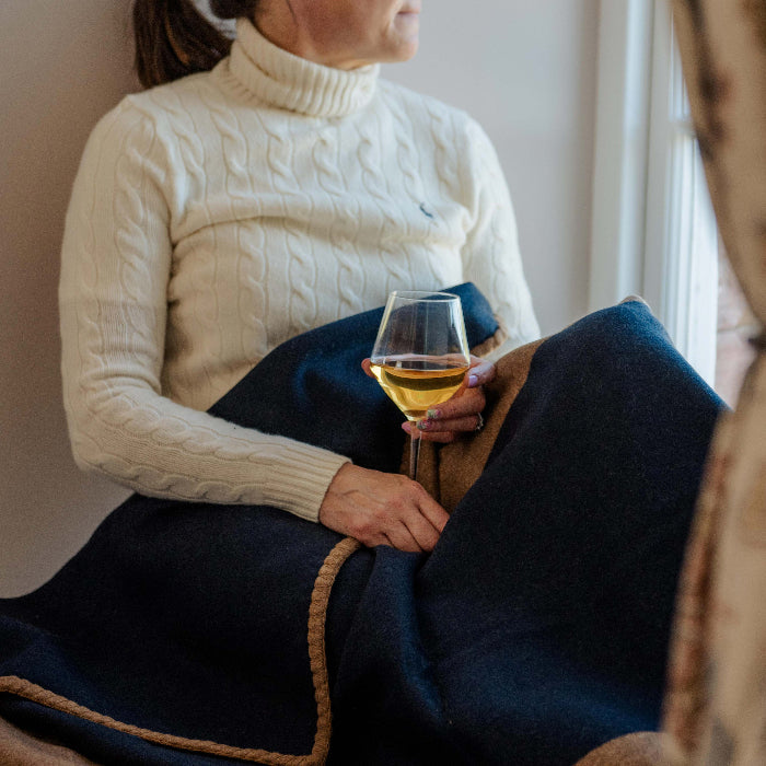 Person sitting by a window holding a glass of wine, with a navy and tan cashmere blanket draped over their legs. 