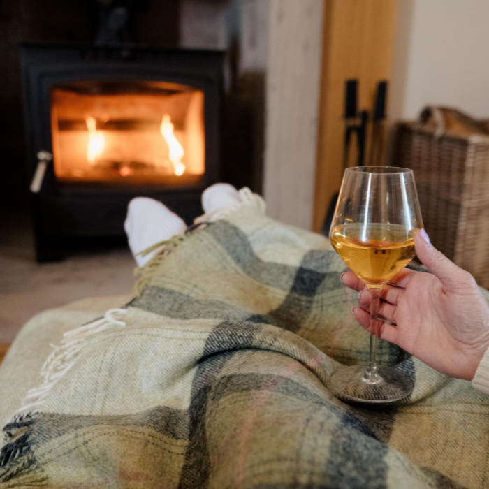 Person holding a glass of wine by a fireplace, wrapped in the Green Skye Check Shetland Wool Throw a  plaid blanket