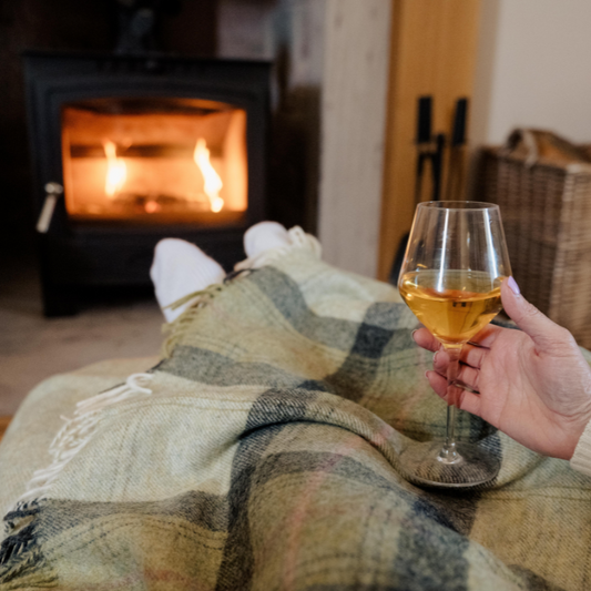 Person holding a glass of wine by a fireplace, wrapped in the Green Skye Check Shetland Wool Throw a  plaid blanket