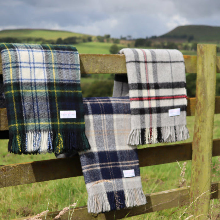 Three plaid blankets including the Grey Thompson, Bannockbane and Dress Gordon Tartan Pure New Wool Blanket hanging on a wooden fence with a scenic background of hills and green fields.