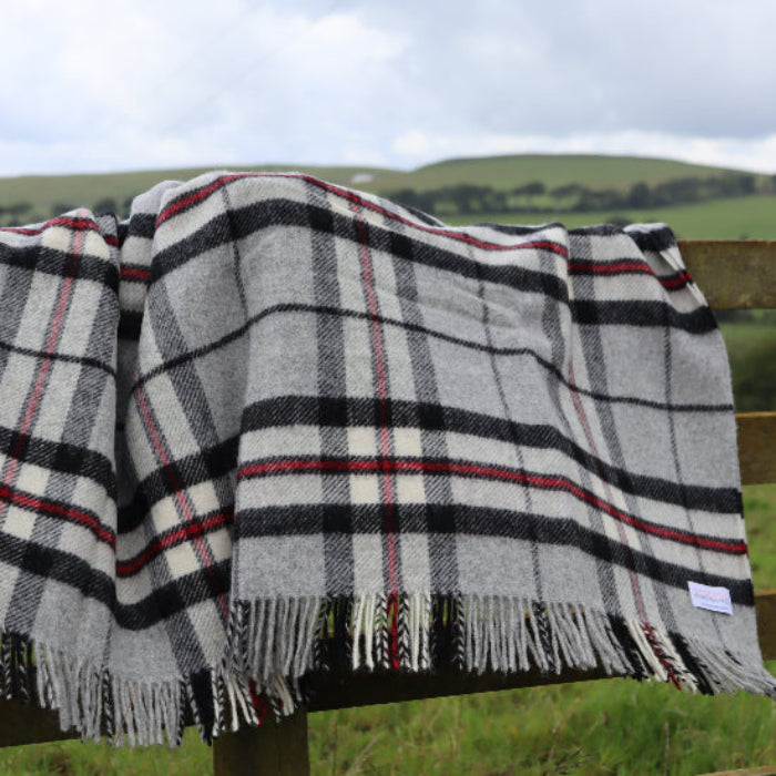 Grey Thompson Tartan Pure New Wool Blanket Rug draped over a wooden fence with a scenic background of green fields and hills.