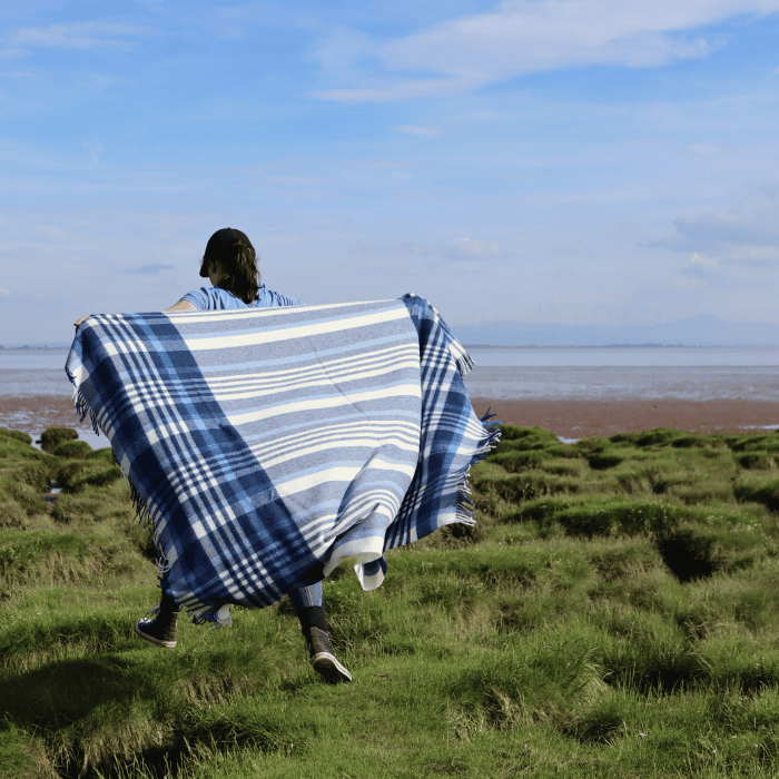Person with a blue and white checked blanket throw walking on a grassy field with a beach in the background.