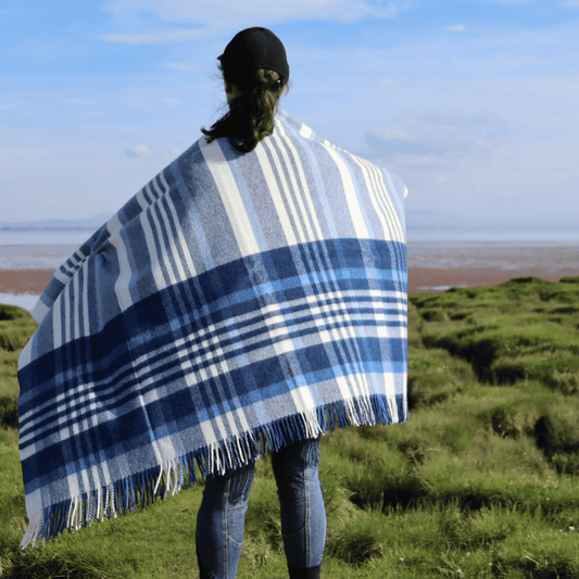 Person wearing a blue checkered lambswool throw standing on a grassy field with a scenic background.