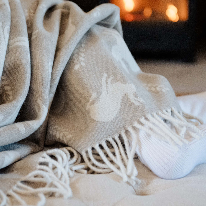 Person wearing white socks sitting on a couch wrapped up in a cosy beige blanket with hares printed on it featuring a fringe in front of a fireplace.