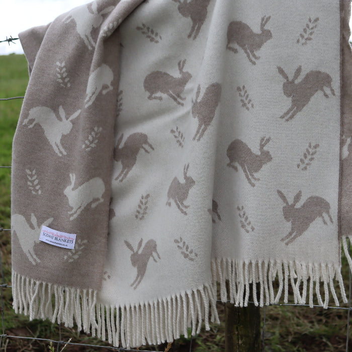 Cosy blanket with hare pattern with a Solway Blankets label draped over a fence in a grassy field
