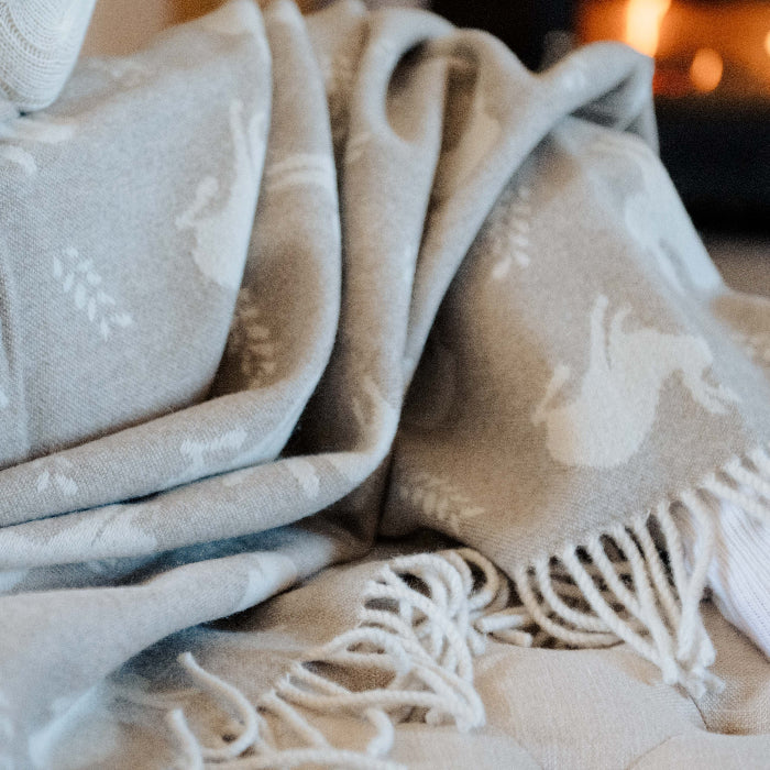 Close up of a cosy beige blanket with hare patterns draped over a person sitting on a couch, with a warm fireplace glow in the background.