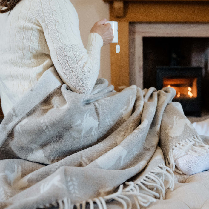 Person sitting on a couch with a cosy blanket with hares printed on it holding a mug in front of a fireplace