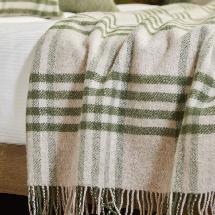 Plaid olive green and beige blanket  draped over the side of a white bed. 