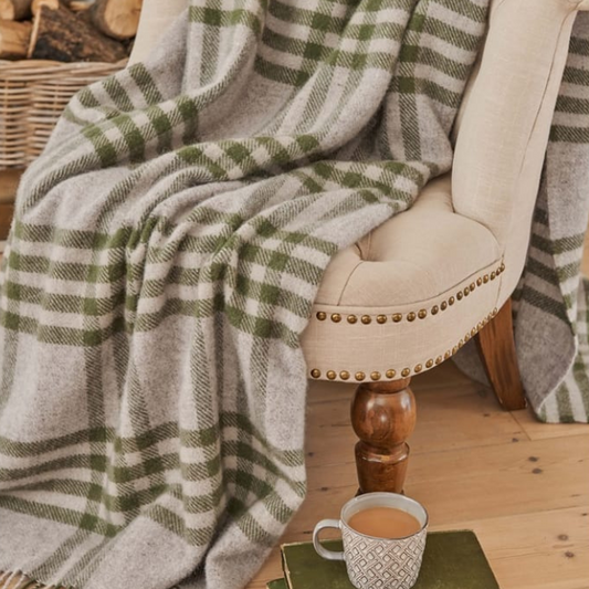 An extra large blanket with a deep olive green and beige check pattern, draped over a beige chair with a cup of coffee on the wooden floor underneath