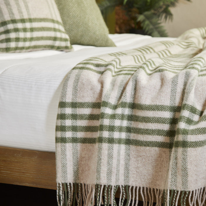 Plaid olive green and beige wool blanket draped over a bed with matching green and beige pillows and a plant in the background. 