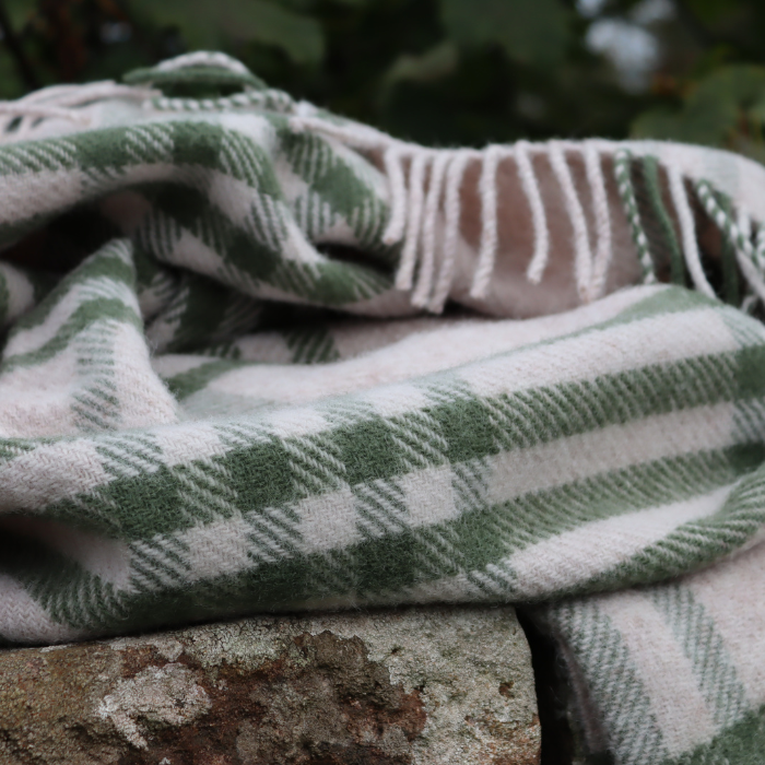Close up of a soft olive green and beige checked wool blanket with fringe, draped outdoors on a mossy stone wall – natural, cosy and crafted in Britain.