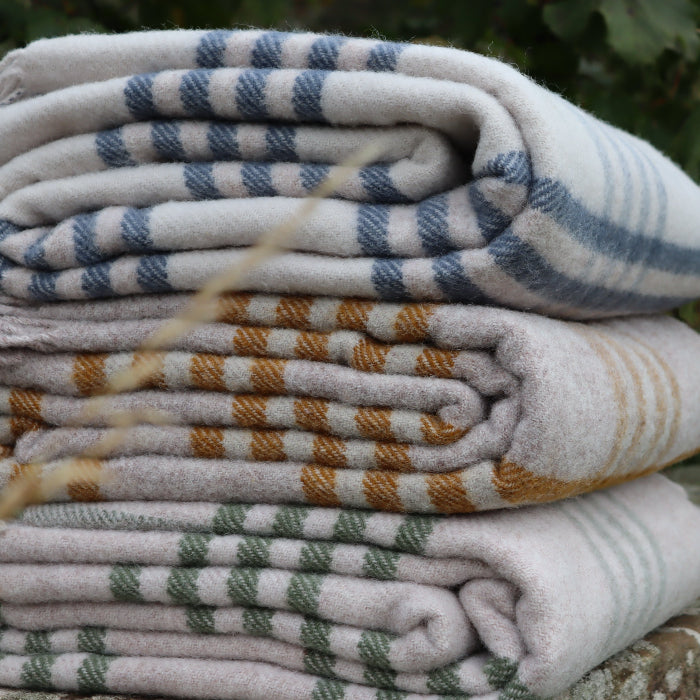Stack of folded wool blankets featuring colours of blue, green and mustard with plaid patterns on a stone surface with greenery in the background