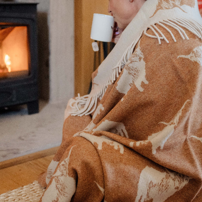 Person wrapped up in a orange lambswool blanket with printed Highland Cows sitting by a cosy fireplace